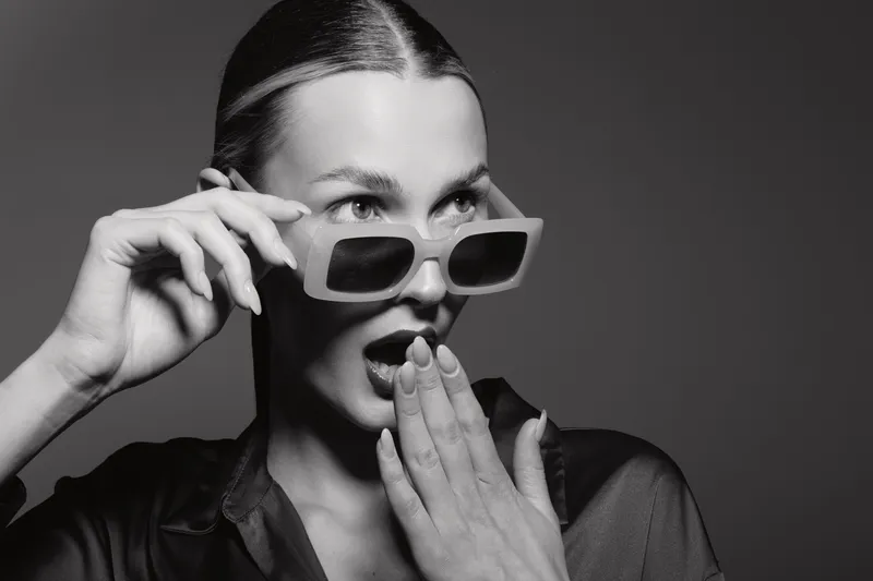 Surprised white woman with light skin and slick hair adjusts sunglasses, covering mouth with hand in studio. Concept of fashion, expression, and modern style.