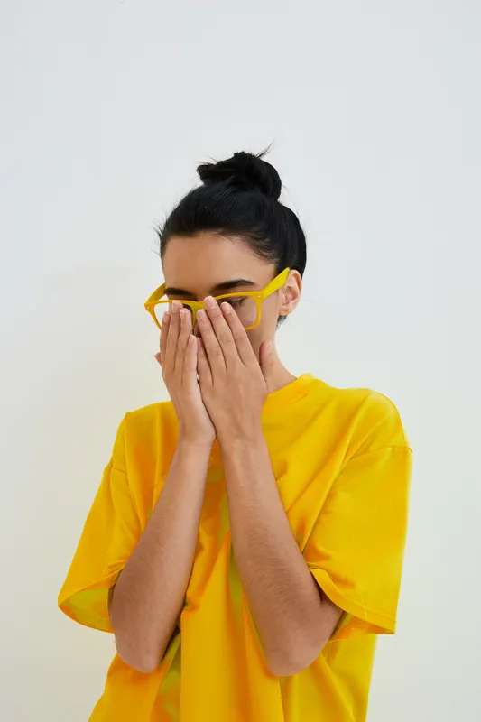Young woman in yellow glasses and t-shirt covers her face with hands, displaying shyness and modesty against a plain white background in casual style.