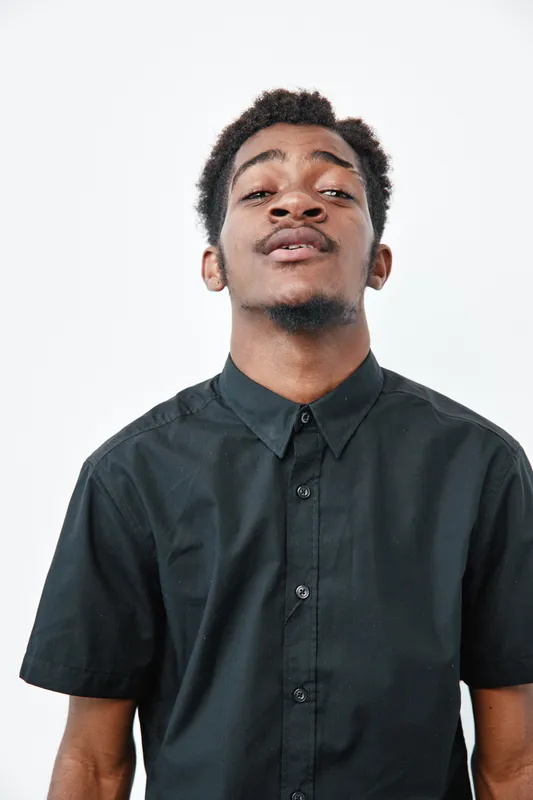 Young black man with dark skin wearing black shirt standing against white background looking confident and casual in studio portrait with natural hair and beard.