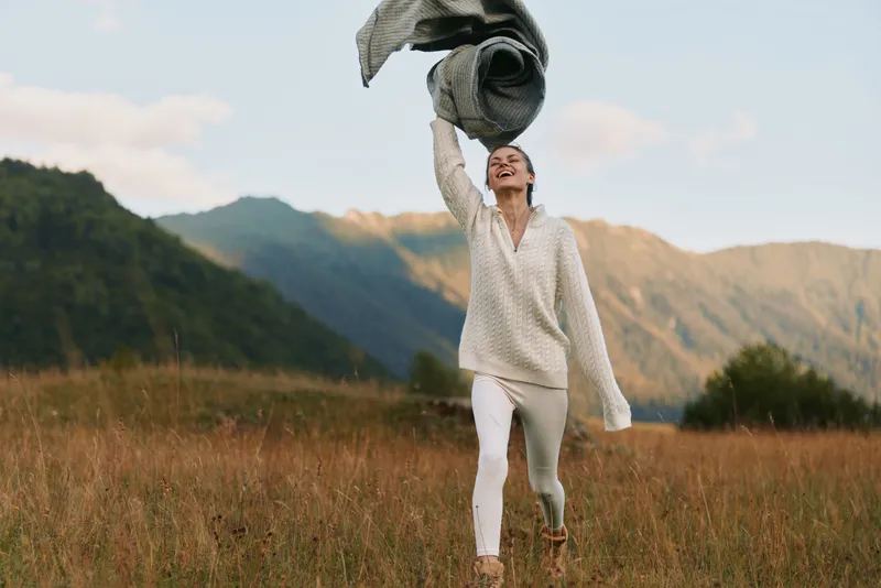 A smiling woman in a white knit sweater and pants runs through a grassy meadow, throwing a scarf into the air with mountains in the background, joyful energy.