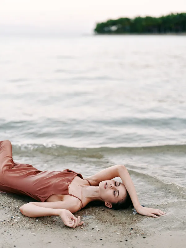 A tranquil beach scene shows a relaxed woman in a soft pink dress lying on the sand, basking in warm light as calm waves lap the shore at sunset.