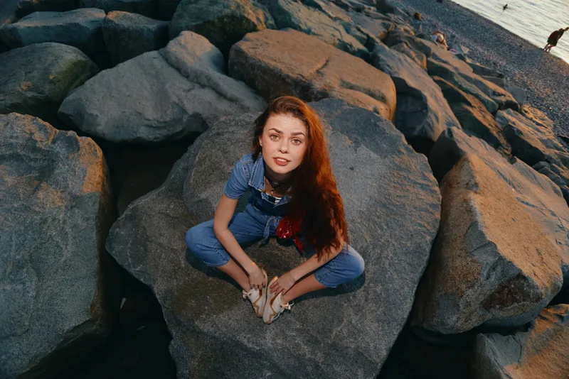 lifestyle woman in modern boho-western denim street style sitting on rocks at sunset, film color with warm tones and casual relaxed mood