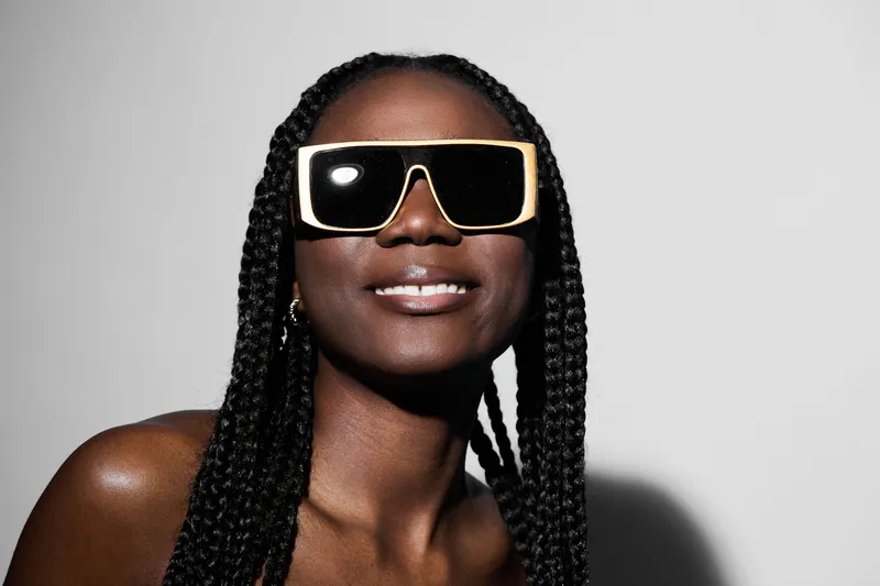 Confident woman with braided hair wearing large sunglasses, smiling against a minimalist background, capturing summer vibes and fashion style