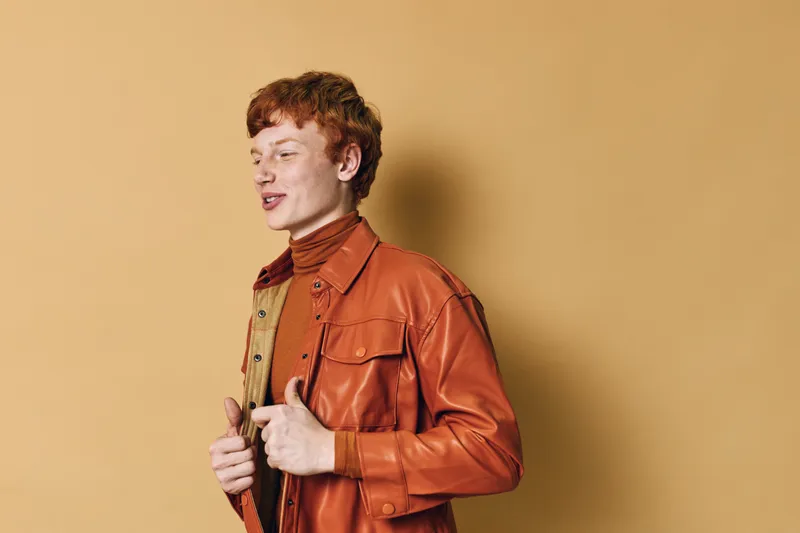 Young redhead man wearing orange leather jacket and turtleneck standing on beige background, posing with confident expression in casual style fashion portrait.