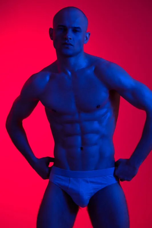 Athletic man posing confidently with a strong physique, illuminated by vibrant blue and red lights, showcasing contrasting colors in a bold and striking manner.