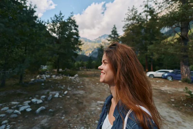 A cheerful woman outdoors in a mountain landscape, smiling and looking to the side. She wears casual denim jacket and white top, enjoying a tranquil natural setting.