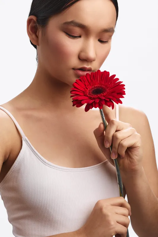 asian woman softly holds a bright red gerbera flower near her face, gazing downward with a calm expression, creating a minimalist portrait that exudes gentle elegance and natural beauty.