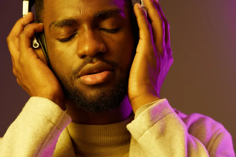 Young Black man enjoying music with headphones, displaying relaxation and happiness, set against a colorful purple and orange background, perfect for music and lifestyle concepts