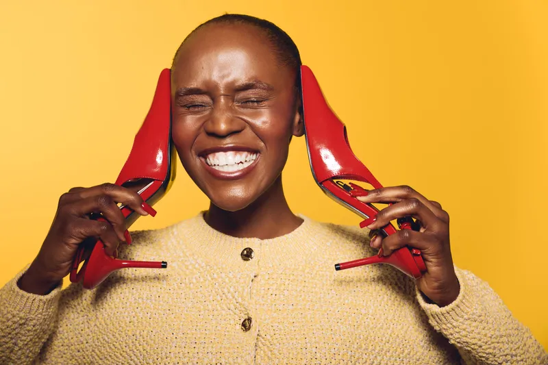 Happy African woman with dark skin laughs holding red high heels near her face on yellow background. Concept for joy, fashion, lifestyle, and beauty with vibrant colors.