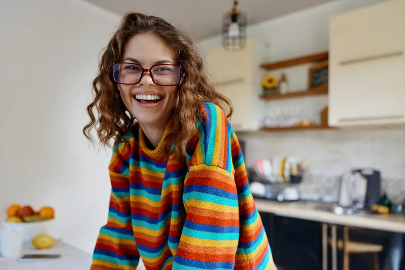 Cheerful woman in a colorful striped sweater smiles in a bright kitchen, capturing a casual home lifestyle scene, a relaxed candid moment with warm light and cozy counters.