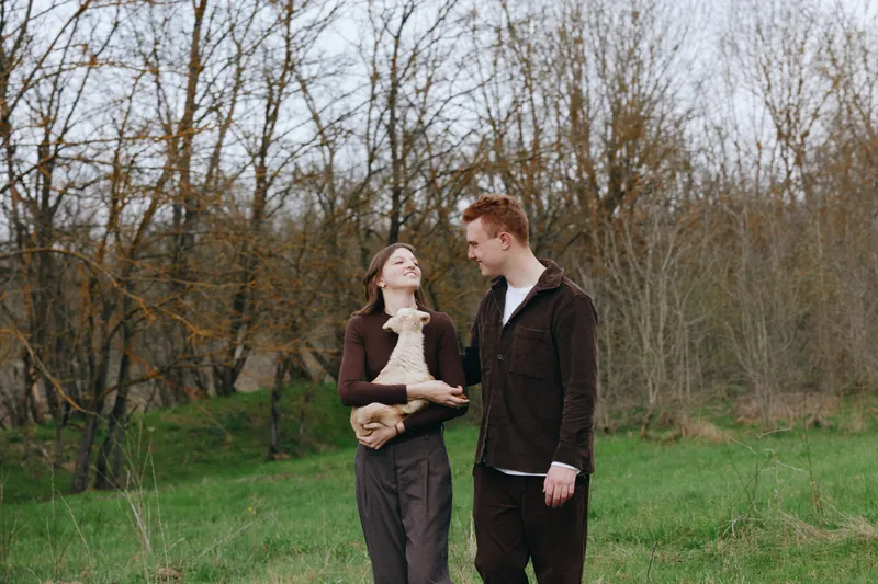 Cheerful couple outdoors, smiling at each other with a baby goat between them, warm sun, casual attire, grassy field, trees in background, light breeze, natural colors