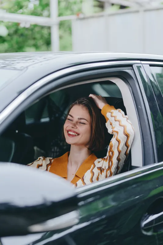 Smiling woman sits in car, relaxed pose with hand through window, wearing an orange striped sweater.