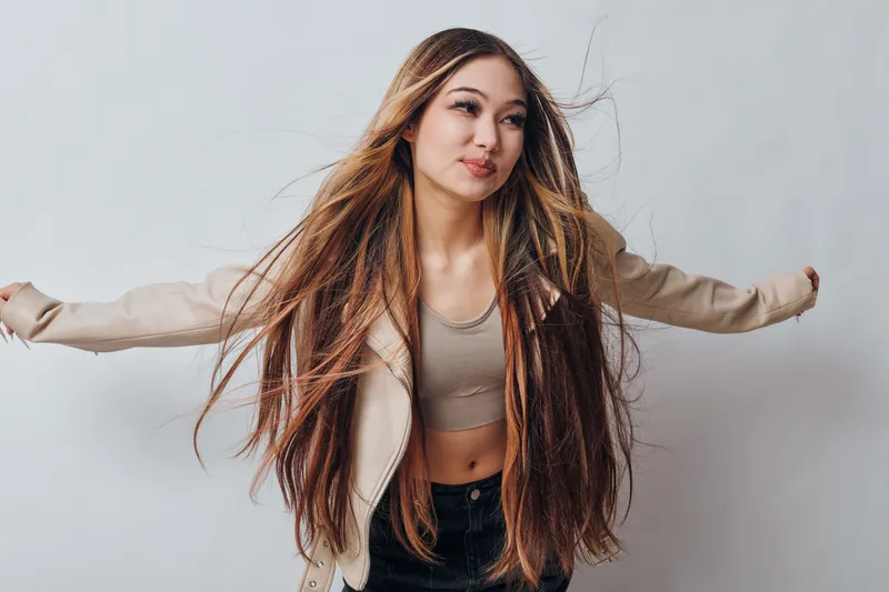 Young woman with long hair wearing casual clothing poses with arms outstretched on light background, expressing freedom confidence and style in studio portrait.