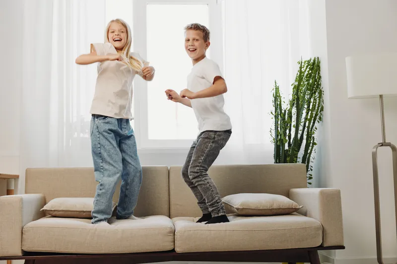 Two happy children jump on a beige sofa in a bright room with natural light, expressing joy and playful energy at home environment.
