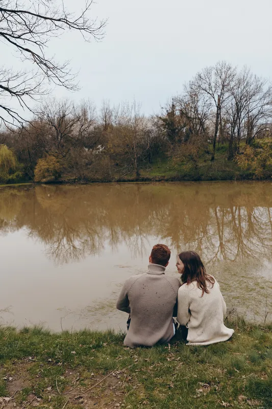 A couple sits on the grassy bank beside a calm lake in autumn, wearing warm sweaters, sharing a quiet moment. A reflective mood and simple lifestyle outdoors.