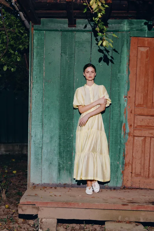 Woman in a pale yellow dress stands on a wooden porch beside a green wall, a calm portrait in natural light, conveying quiet elegance and outdoor rustic charm