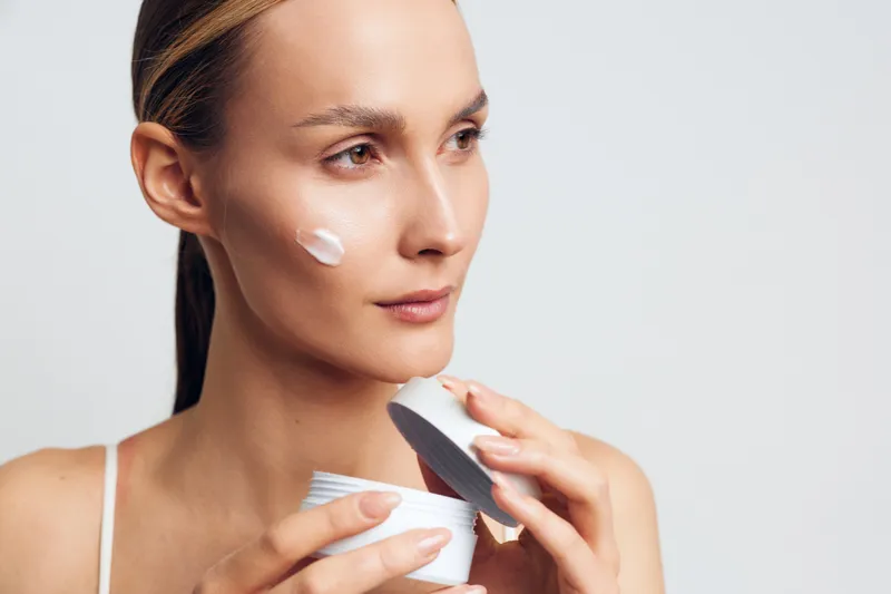 Young woman applying face cream on cheek, clear skin, focused expression, skincare routine, natural light, close-up portrait, minimal makeup, white background