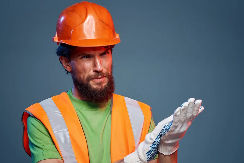 Профессиональный обрезанный портрет бородатого мужчины в оранжевой каске и перчатках на синем фоне