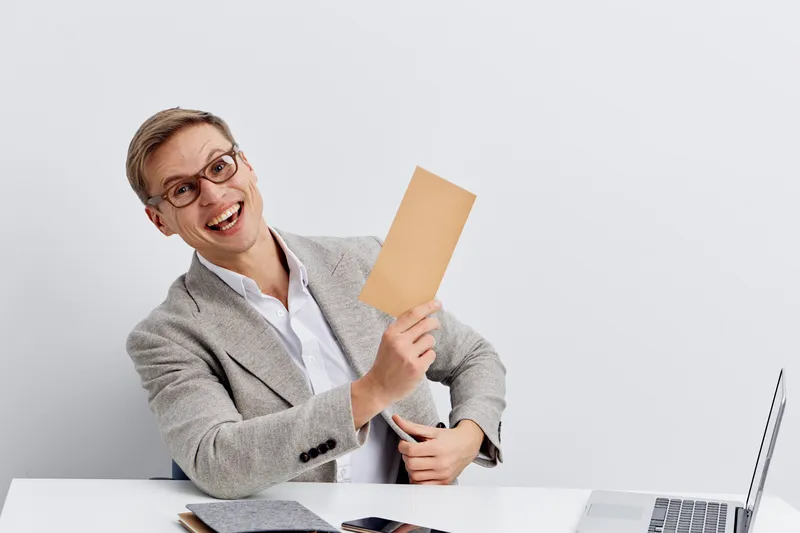 Cheerful woman with short gray hair, glasses, and light gray blazer holding blank card, sitting at desk with laptop and notebook, isolated on plain white background, studio portrait. Business communic