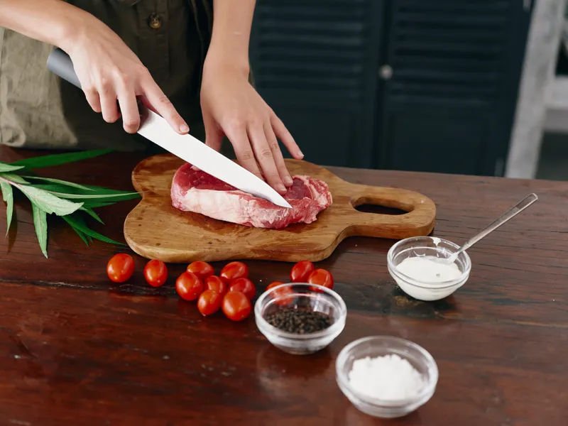 Женщина с ножом в руке нарезает свежее стейковое мясо для запекания на кухне