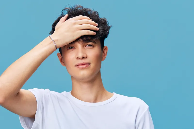 young man with dark curly hair wearing white t-shirt touching forehead against blue background, casual look with slight smile and bracelet accessory