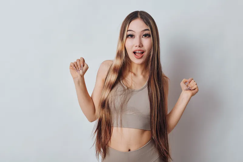 Young woman with long braided hair wearing sportswear showing excited gesture with raised fists and open mouth against plain white background.