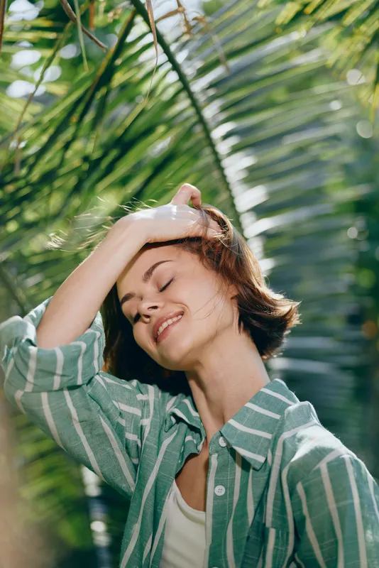 A woman smiles with eyes closed under bright sunlight, wearing a green striped shirt, in a lush outdoor setting.