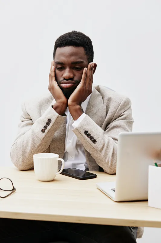 Thoughtful young man of African descent in beige blazer relaxing at workspace, dedicating attention to computer, with coffee cup and smartphone on light wood desk, neutral background, indoor business 
