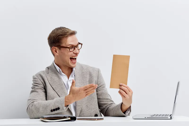 Cheerful young man wearing glasses and stylish gray blazer holding a brown envelope while sitting at a desk with a laptop isolated on white background studio portrait. People lifestyle concept