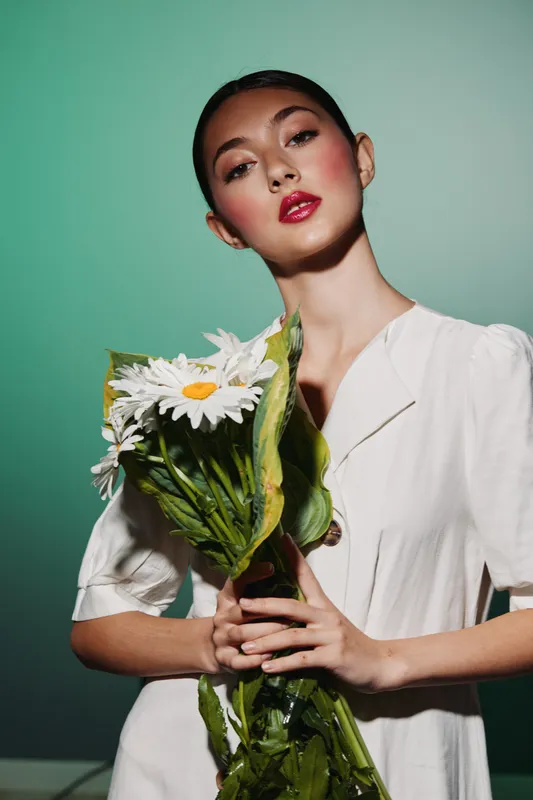 Young woman with red lips holding white daisies wears a stylish minimal white outfit. Green gradient background enhances beauty and fashion concept with floral elements.