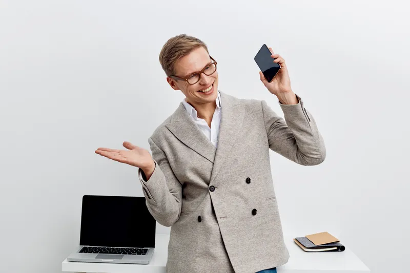 smiling man communicating video call holding smartphone wearing gray coat standing near laptop in modern office remote work digital nomad concept