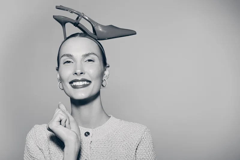 Smiling Caucasian woman with fair skin holding high heel shoe on head in white sweater. Concept of fashion, fun, and creative portrait.