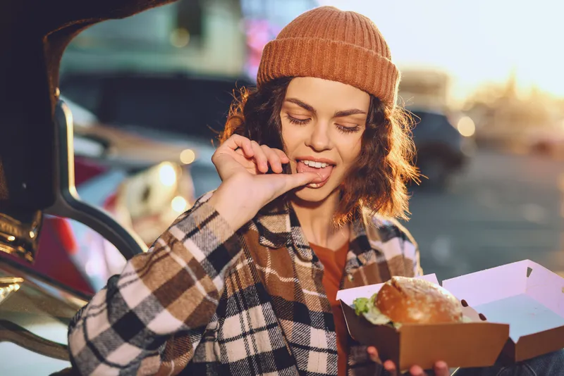woman, burger, takeout, car, beanie, smile - candid lifestyle moment of a young woman eating a takeout burger by her car at golden hour glow, authenticity and emotional storytelling capture joyful