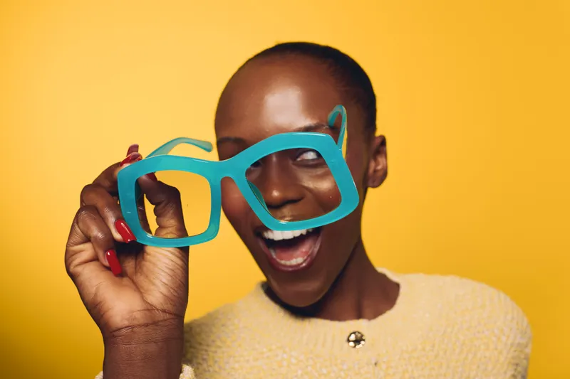 Happy dark skinned African woman holding oversized blue glasses near her face on yellow background. Concept of joy, fashion, fun, and playful emotions in studio portrait.