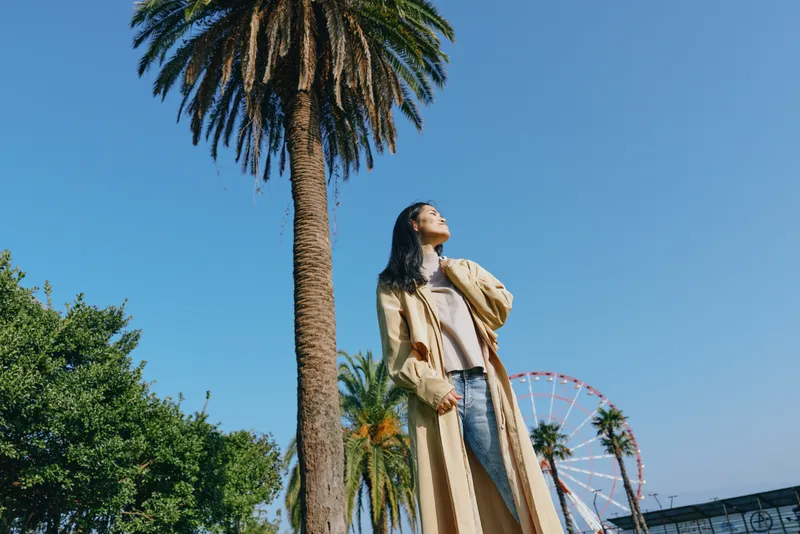 Woman standing beneath a tall palm tree in a sunny park, overlooking a distant Ferris wheel, wearing a long coat and jeans, capturing a carefree summer moment.