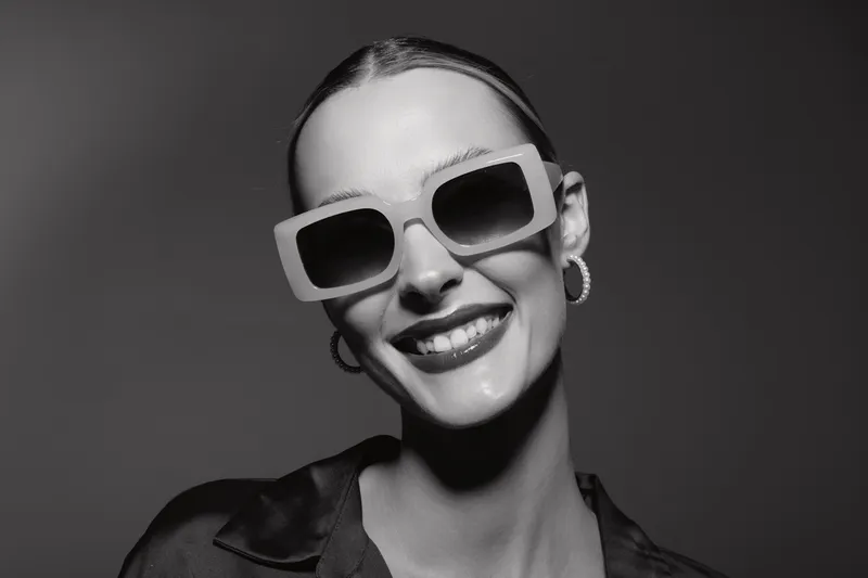 Smiling Caucasian woman with pale skin wearing oversized sunglasses and hoop earrings. Close-up portrait in black and white with fashionable style and confident expression.