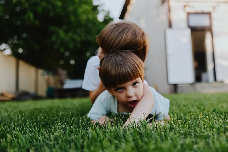 Playful children enjoying a sunny day in a backyard, one boy lying down while the other playfully hugs him from behind, surrounded by lush green grass.