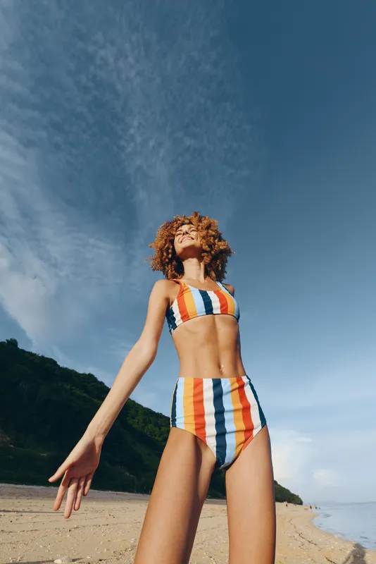 Beach scene, woman in striped swimsuit, curly hair, ocean breeze, summer vacation feeling, she stands on sand with arms open, blue sky and clouds, outdoor lifestyle