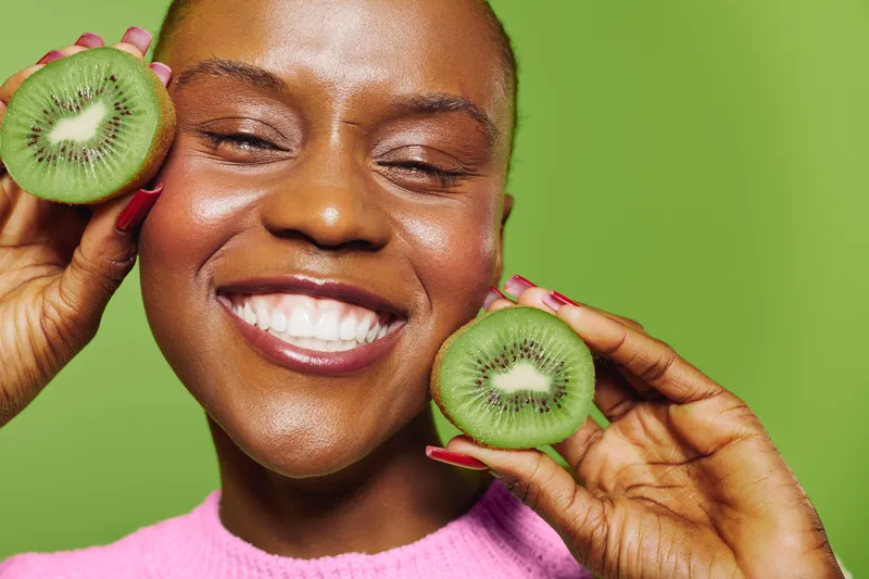 Happy black woman with dark skin smiles broadly while holding two halves of fresh kiwi fruit near her face. Bright green background and pink sweater create a vibrant, healthy lifestyle concept.