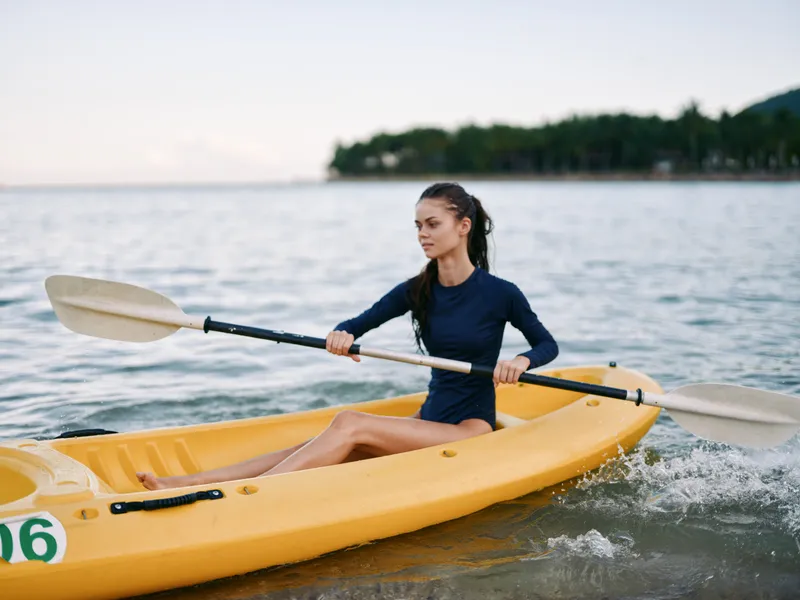 Main subject is a young woman paddling a yellow kayak across calm water, using a double blade paddle while wearing a navy long sleeve top and focused expression.