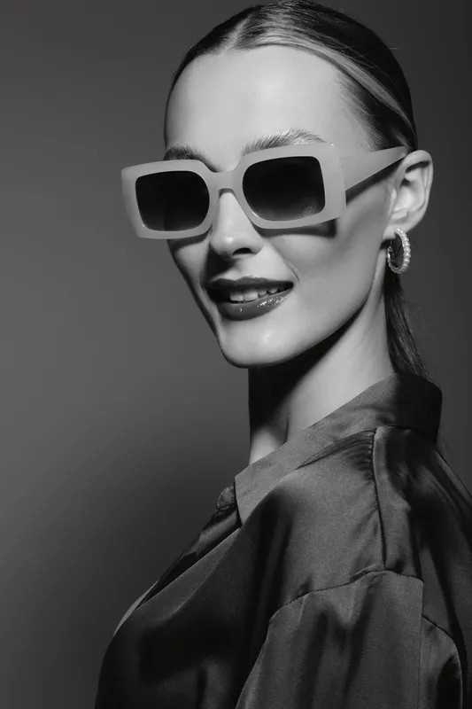 Caucasian woman with fair skin wearing large sunglasses and silk blouse smiles confidently in studio with dramatic lighting. Concept of fashion, elegance, and style.