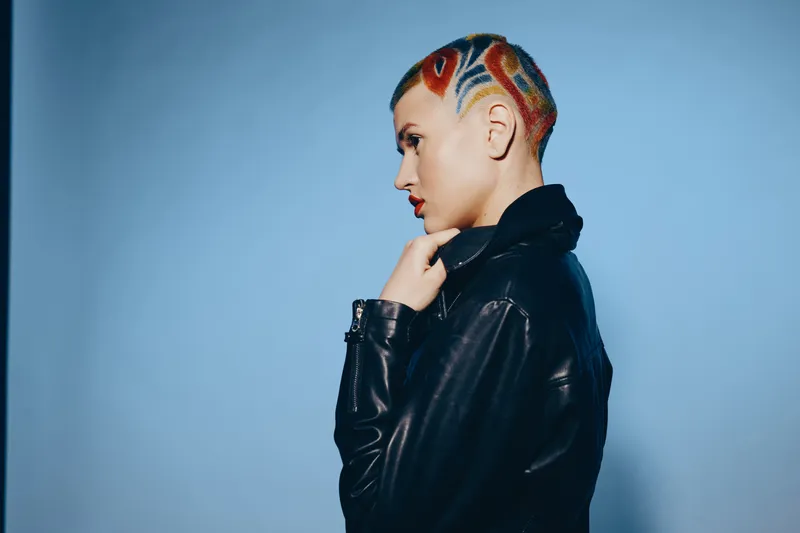 woman with colorful geometric shaved hairstyle wearing black leather jacket stands against soft blue background in dramatic side profile exhibiting bold modern style