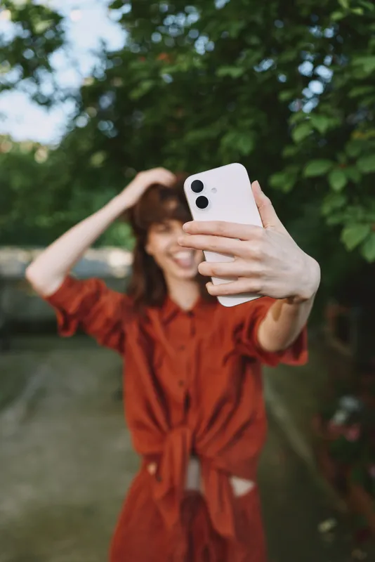Woman in a red dress takes a cheerful selfie outdoors among lush greenery, showcasing a casual lifestyle moment in a sunny park setting, smiling at the camera