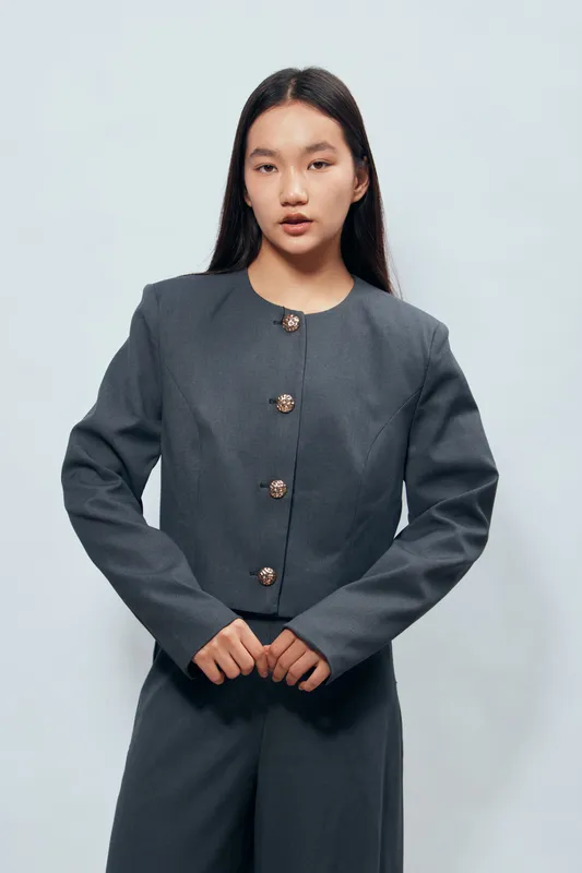Young Asian woman wearing a tailored grey jacket and matching pants standing with hands together in front against light background. Fashion model posing with natural expression and long hair.