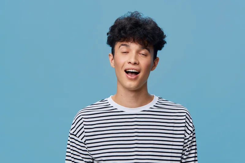 smiling young man with curly hair wearing striped shirt standing against blue background with eyes closed, expressing happiness and relaxation.