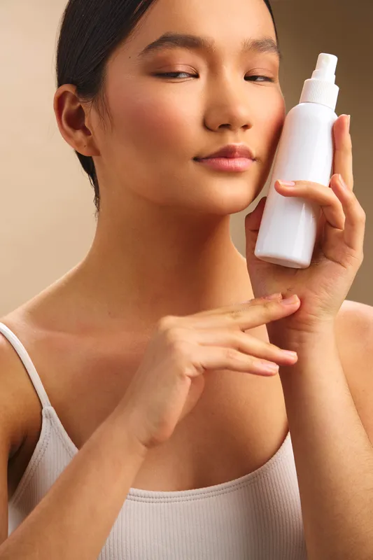Asian woman applies skincare product in a calm home setting, showcasing clear skin and gentle care. The soft light highlights healthy complexion, conveying wellness, beauty, and self care through a