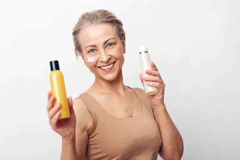 Smiling mature woman with skincare patches under eyes holds two cosmetic bottles, showcasing beauty and self-care routine on white background. Concept of wellness and facial treatment.