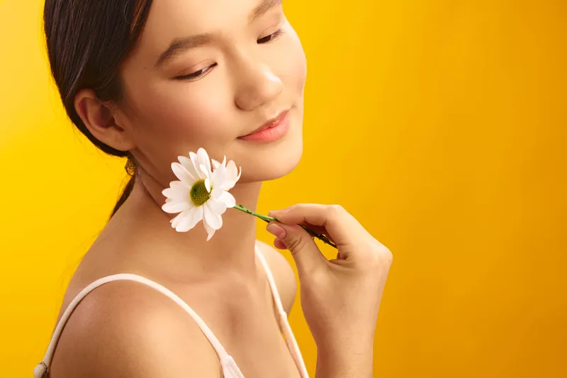 asian woman gracefully holds a white flower near her face against a bright yellow backdrop, exuding calm elegance and soft beauty with a gentle smile and serene expression, inviting warmth and