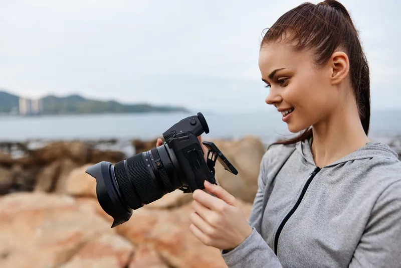 A smiling young woman checking her camera on a rocky beach, wearing a grey hoodie, reflecting a casual and passionate approach to photography by the sea