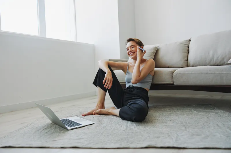 Фрилансерка с ноутбуком и телефоном работает из дома, сидя на полу в домашней одежде, с короткой стрижкой
